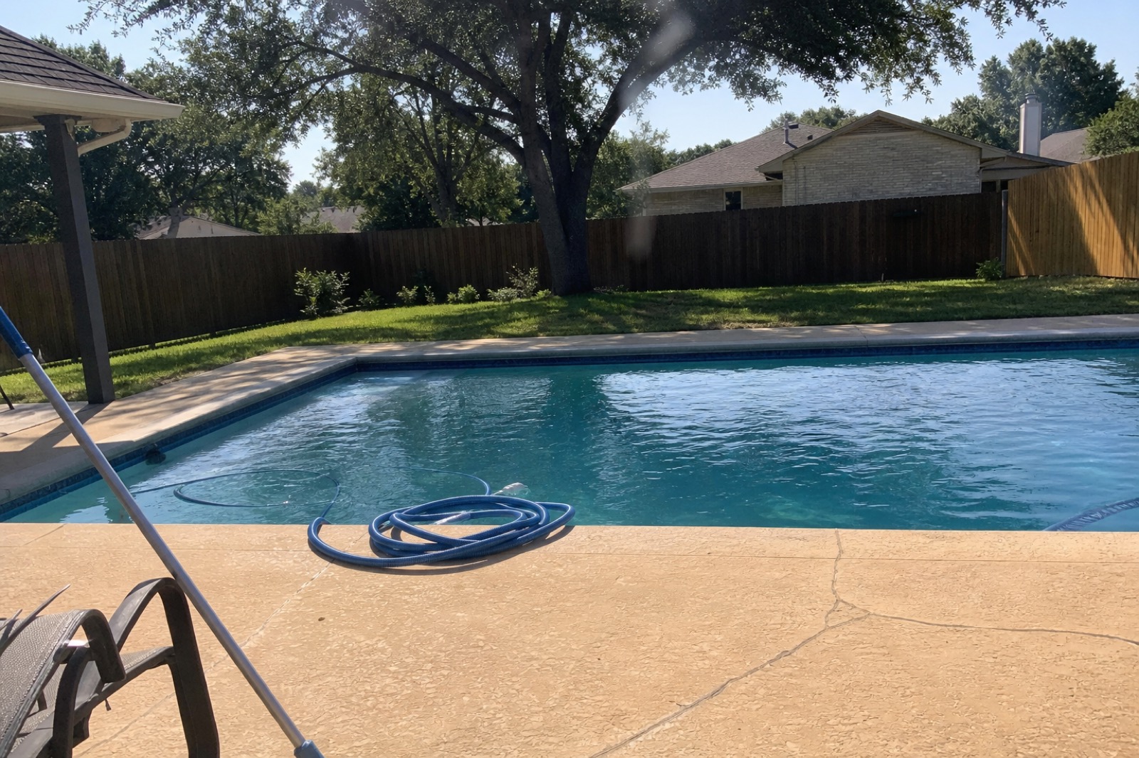 Backyard pool in a Southwest Fort Worth neighborhood at midday, telescoping pole leaning by the deck and a coiled vacuum hose near the skimmer.