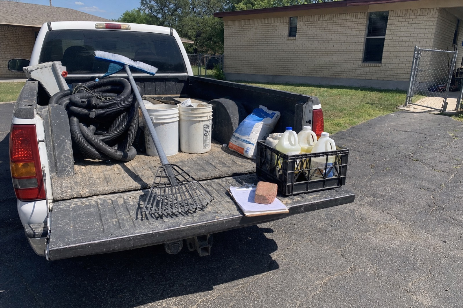 The bed of a work pickup loaded for a weekly pool route: telescoping pole, vacuum hose, five-gallon buckets, salt bag, milk crate with jugs of liquid chlorine and acid, and a clipboard route sheet held down by a brick.